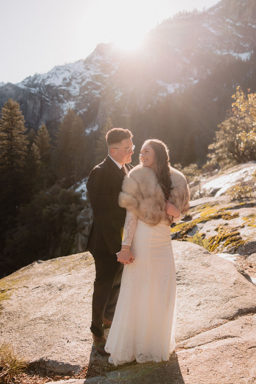 A couple in formal attire stands on a rocky surface with mountains and trees in the background, illuminated by sunlight.