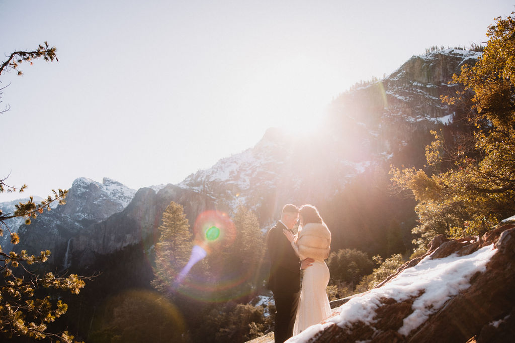 A couple in formal attire stands on a rocky surface with mountains and trees in the background, illuminated by sunlight.