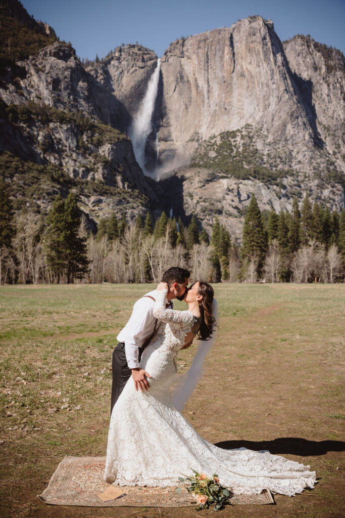 A bride and groom embrace outdoors, with mountains in the background. The bride wears a veil and holds flowers; the groom is in a white shirt with suspenders for an elopement in california