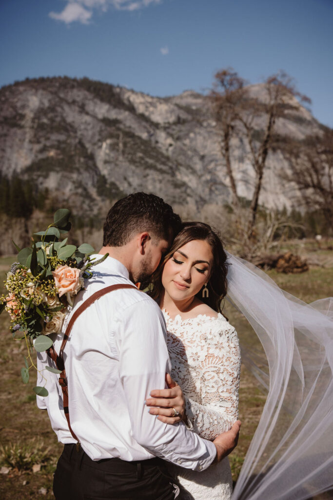 A bride and groom embrace outdoors, with mountains in the background. The bride wears a veil and holds flowers; the groom is in a white shirt with suspenders for an elopement in california