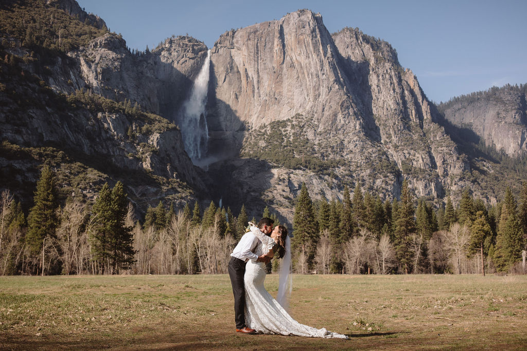 A bride and groom embrace outdoors, with mountains in the background. The bride wears a veil and holds flowers; the groom is in a white shirt with suspenders for an elopement in california