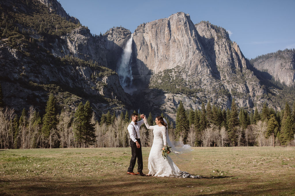 A bride and groom embrace outdoors, with mountains in the background. The bride wears a veil and holds flowers; the groom is in a white shirt with suspenders for an elopement in california