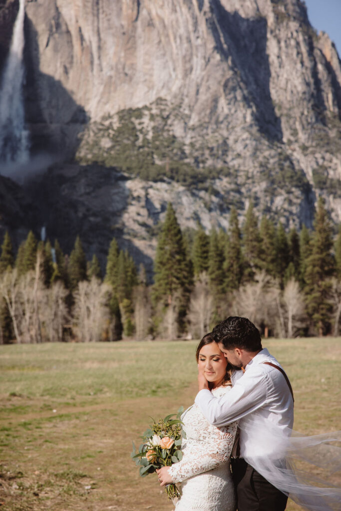 A bride and groom embrace outdoors, with mountains in the background. The bride wears a veil and holds flowers; the groom is in a white shirt with suspenders for an elopement in california