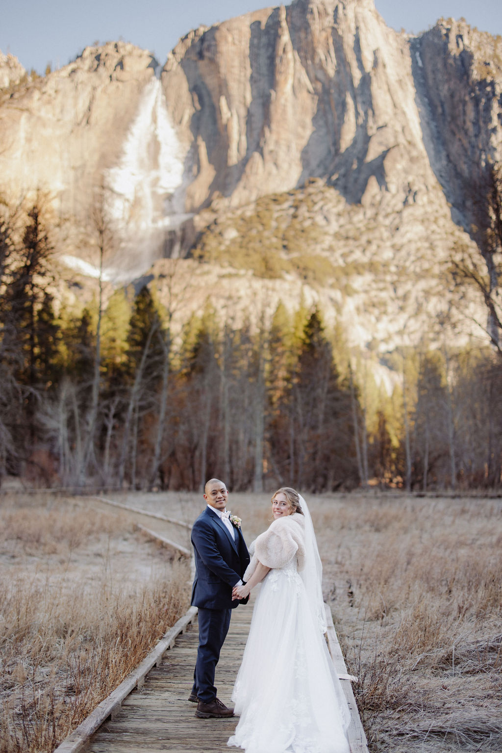 A bride and groom stand on a wooden path in an open field, holding hands, with tall mountains and pine trees in the background.