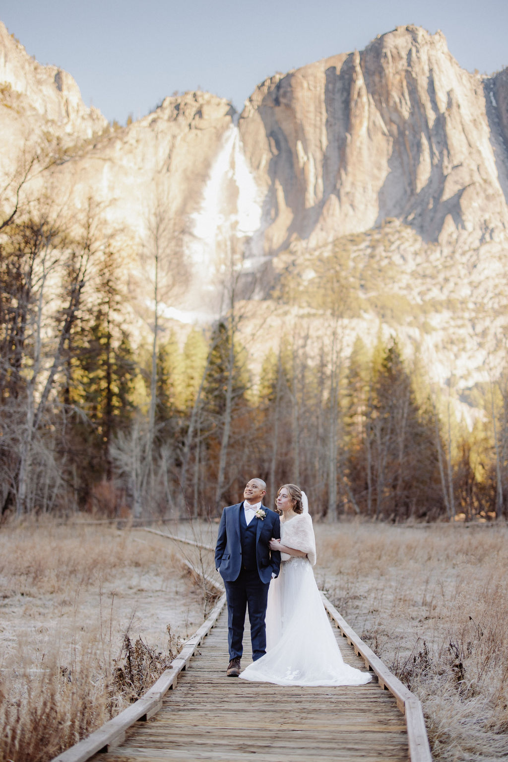 A bride and groom stand on a wooden path in an open field, holding hands, with tall mountains and pine trees in the background.