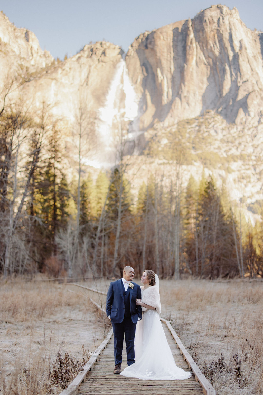 A bride and groom stand on a wooden path in an open field, holding hands, with tall mountains and pine trees in the background.