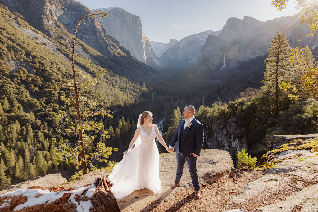 A bride and groom on a rocky ledge with mountains and forest in the background, under daylight for their sunrise yosemite elopement