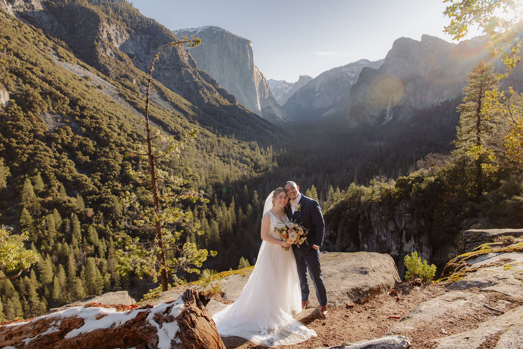 A bride and groom on a rocky ledge with mountains and forest in the background, under daylight for their sunrise yosemite elopement