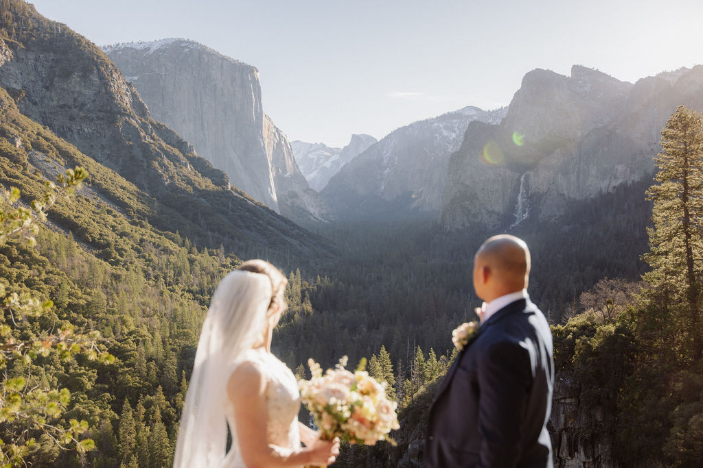 A bride and groom on a rocky ledge with mountains and forest in the background, under daylight for their sunrise yosemite elopement