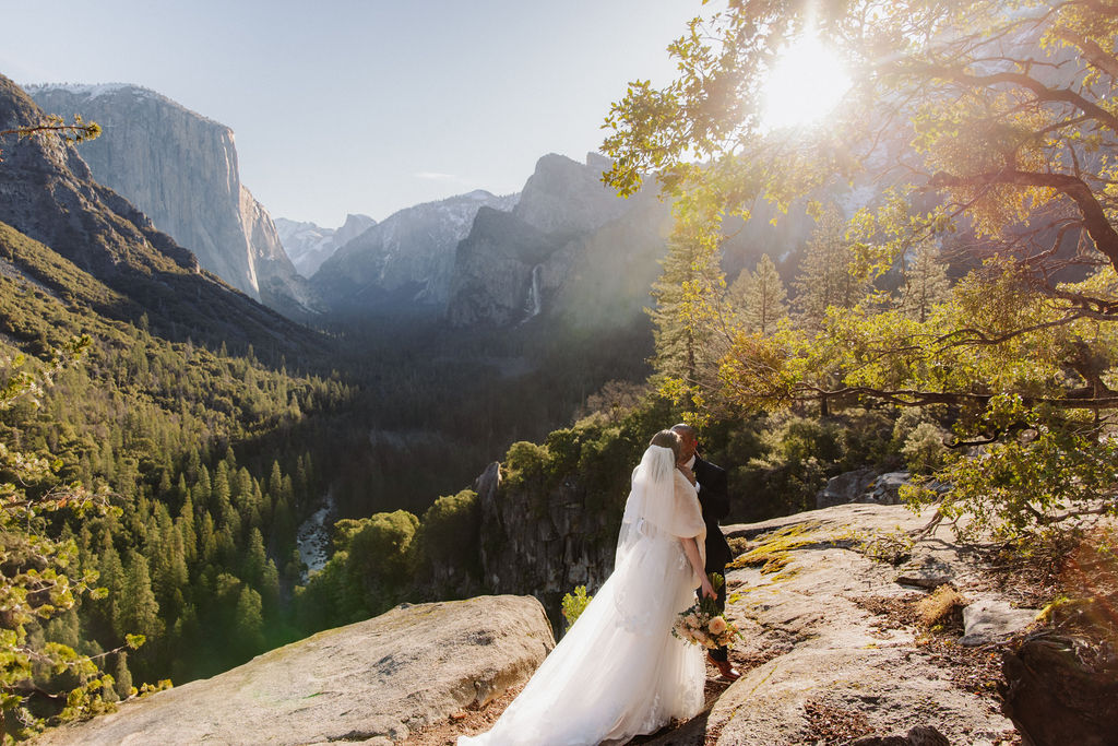 A bride and groom on a rocky ledge with mountains and forest in the background, under daylight for their sunrise yosemite elopement