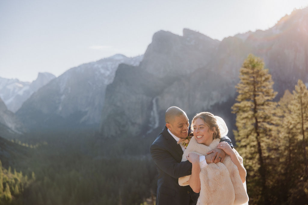A bride and groom on a rocky ledge with mountains and forest in the background, under daylight for their sunrise yosemite elopement