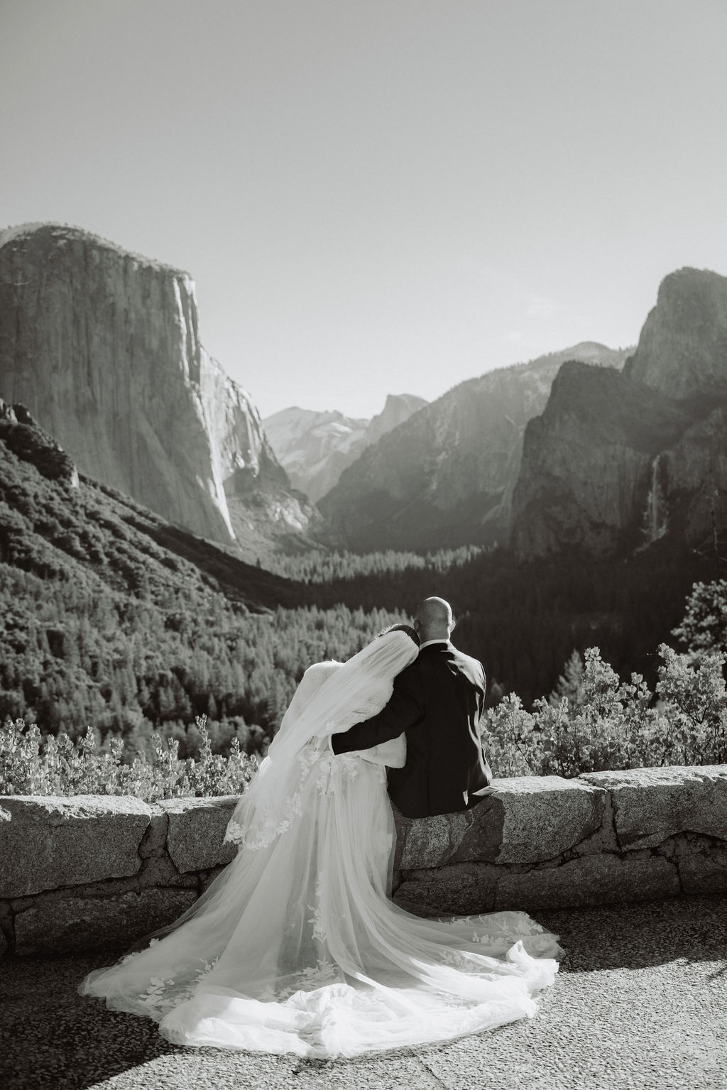 A bride and groom on a rocky ledge with mountains and forest in the background, under daylight for their sunrise yosemite elopement