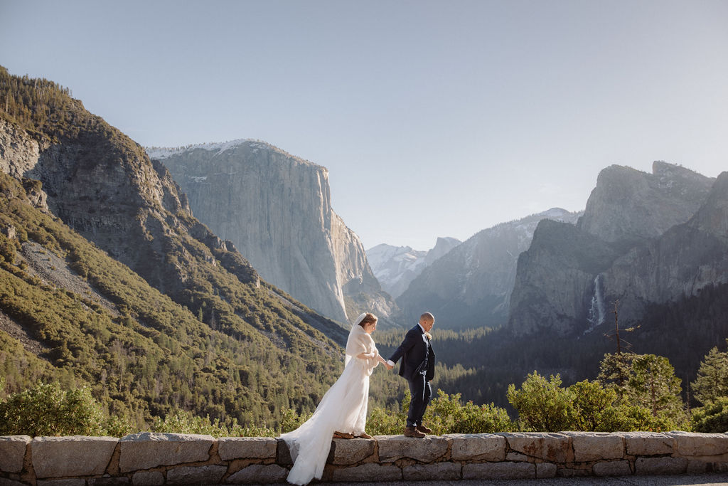 A bride and groom on a rocky ledge with mountains and forest in the background, under daylight for their sunrise yosemite elopement