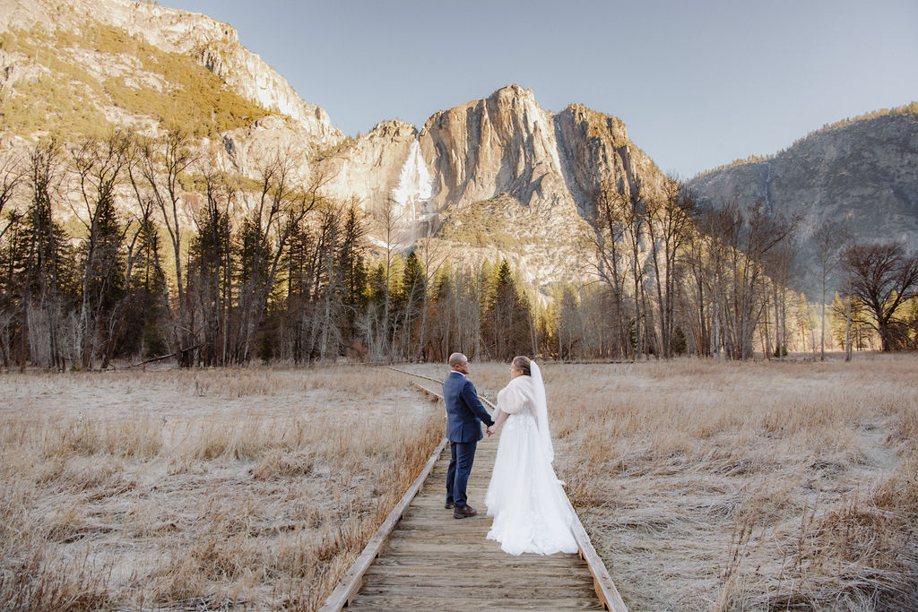 A bride and groom stand on a wooden path in an open field, holding hands, with tall mountains and pine trees in the background.