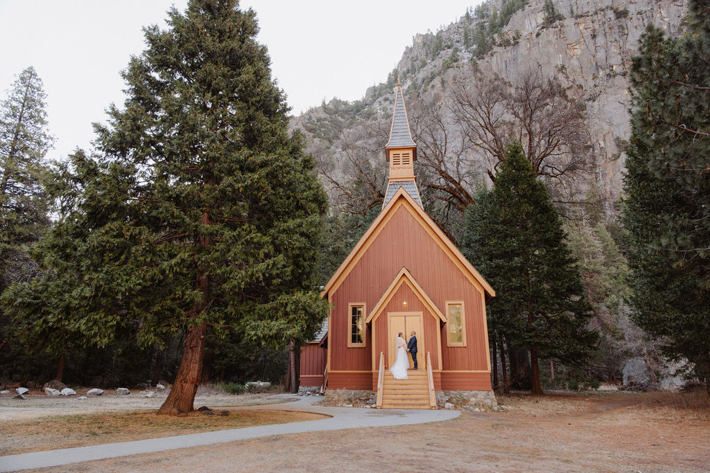 A couple stands at the entrance of a small, red wooden chapel surrounded by tall trees and mountains.