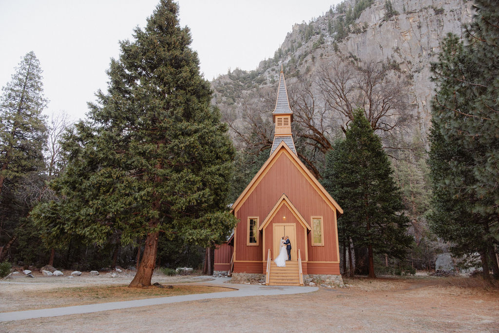 A couple stands at the entrance of a small, red wooden chapel surrounded by tall trees and mountains.