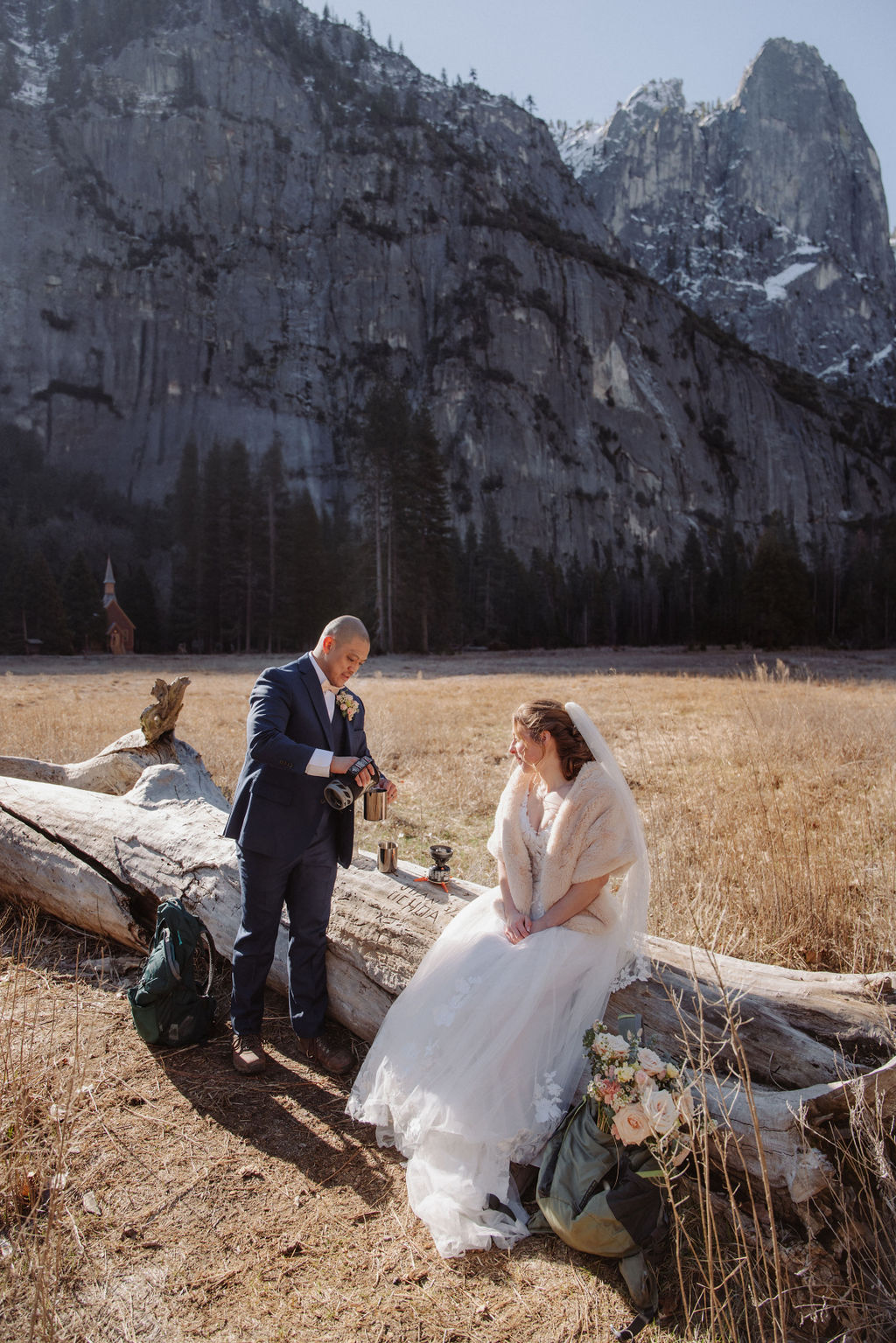 A bride and groom stand on a fallen tree trunk in a grassy field with mountains and a small chapel in the background.