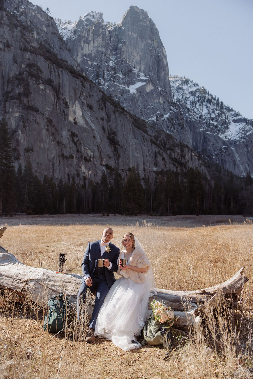 A bride and groom stand on a fallen tree trunk in a grassy field with mountains and a small chapel in the background.