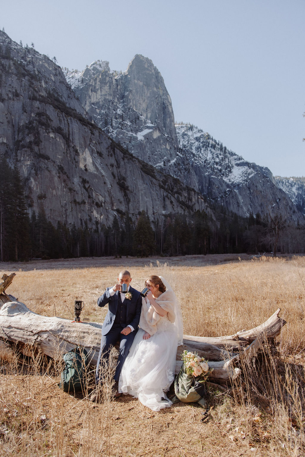 A bride and groom stand on a fallen tree trunk in a grassy field with mountains and a small chapel in the background.