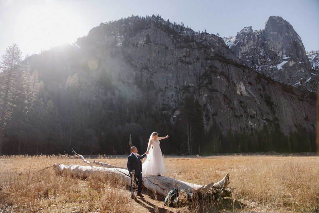 A bride and groom stand on a fallen tree trunk in a grassy field with mountains and a small chapel in the background.