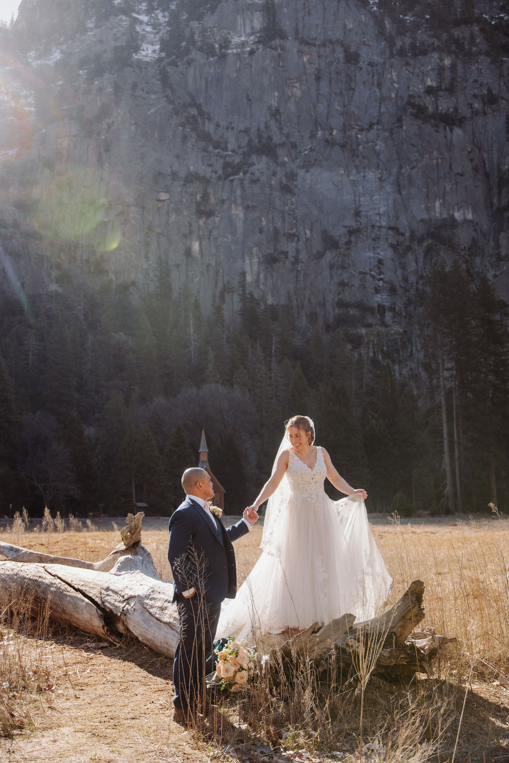 A bride and groom stand on a fallen tree trunk in a grassy field with mountains and a small chapel in the background.