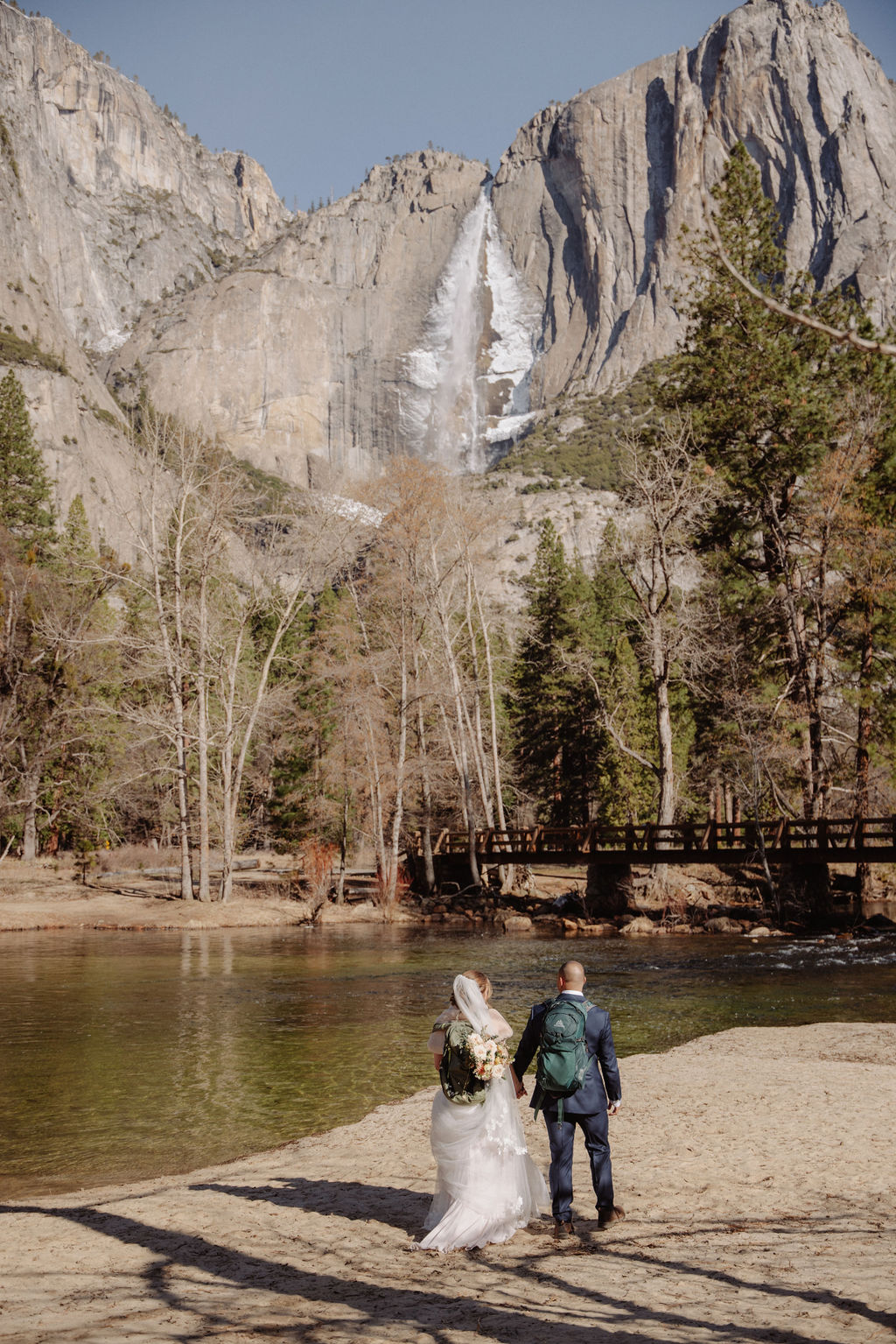 A bride and groom walk along a riverside path, holding hands, with a waterfall and rocky mountains in the background.