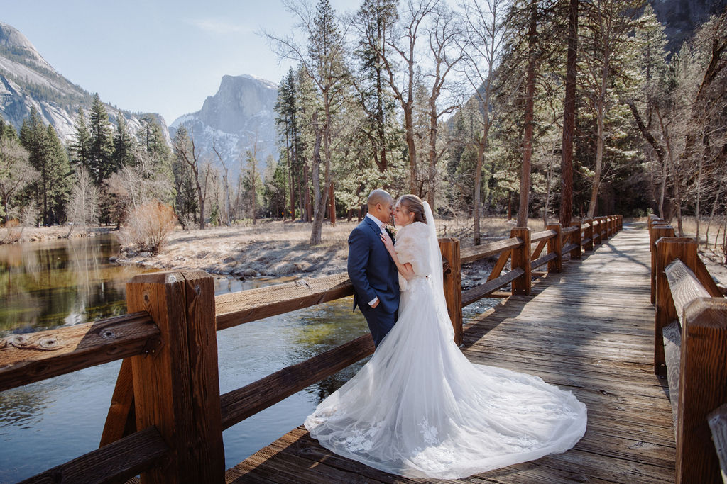 A bride and groom stand close together on a wooden bridge in a forested mountainous area, with trees and a river in the background.