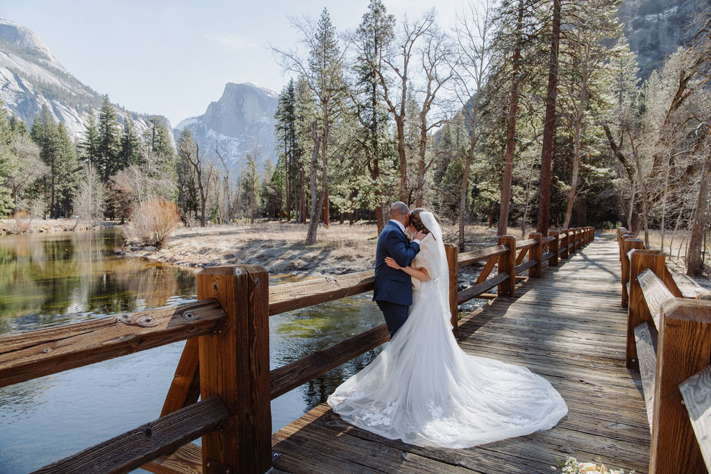 A bride and groom stand close together on a wooden bridge in a forested mountainous area, with trees and a river in the background.