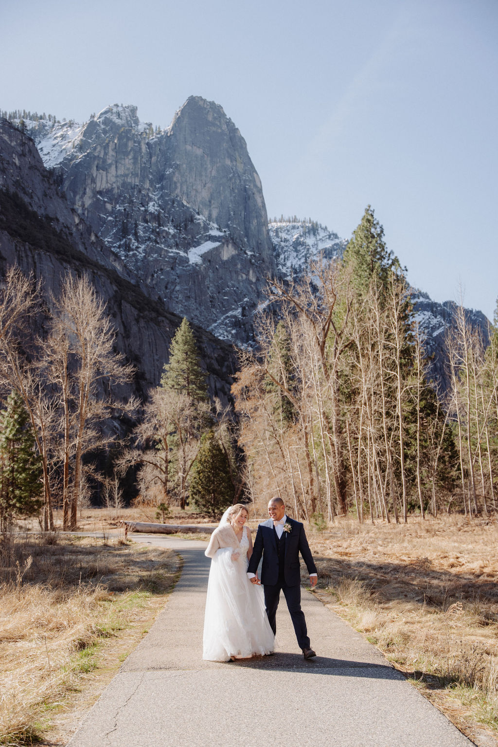 A bride and groom walk hand in hand on a paved path with tall mountains and trees in the background under a clear sky.