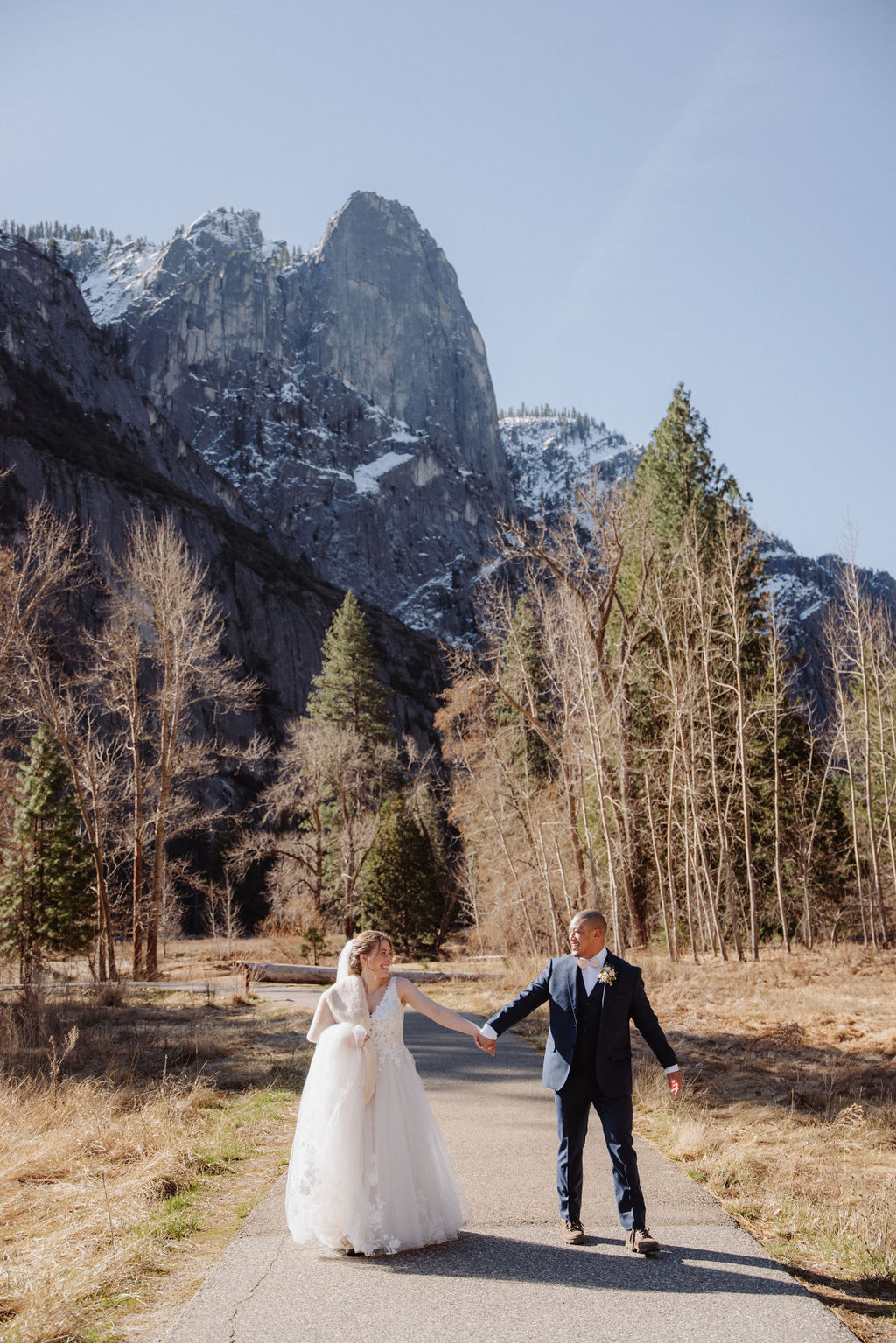 A bride and groom walk hand in hand on a paved path with tall mountains and trees in the background under a clear sky.