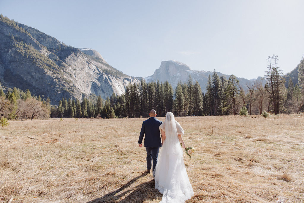 A bride and groom walk hand in hand on a paved path with tall mountains and trees in the background under a clear sky.