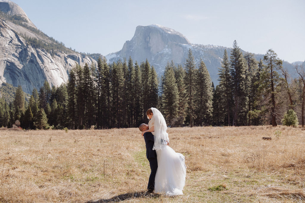 A groom lifts a bride in a wedding dress in a field with pine trees and mountains in the background under a clear sky for a sunrise yosemite elopement