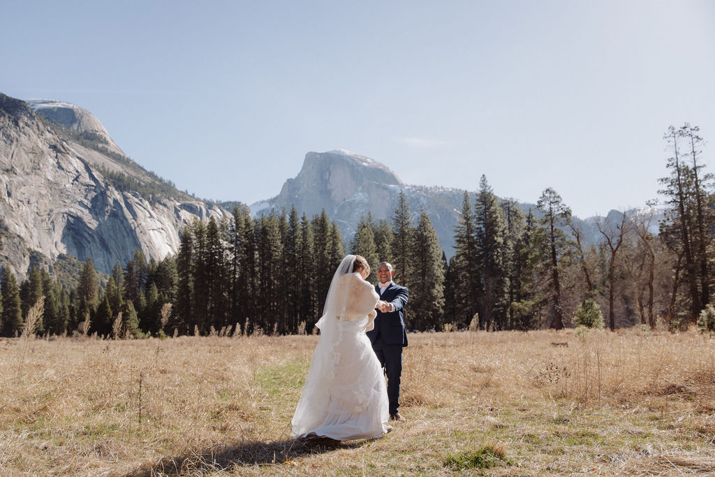 A bride and groom walk hand in hand on a paved path with tall mountains and trees in the background under a clear sky.