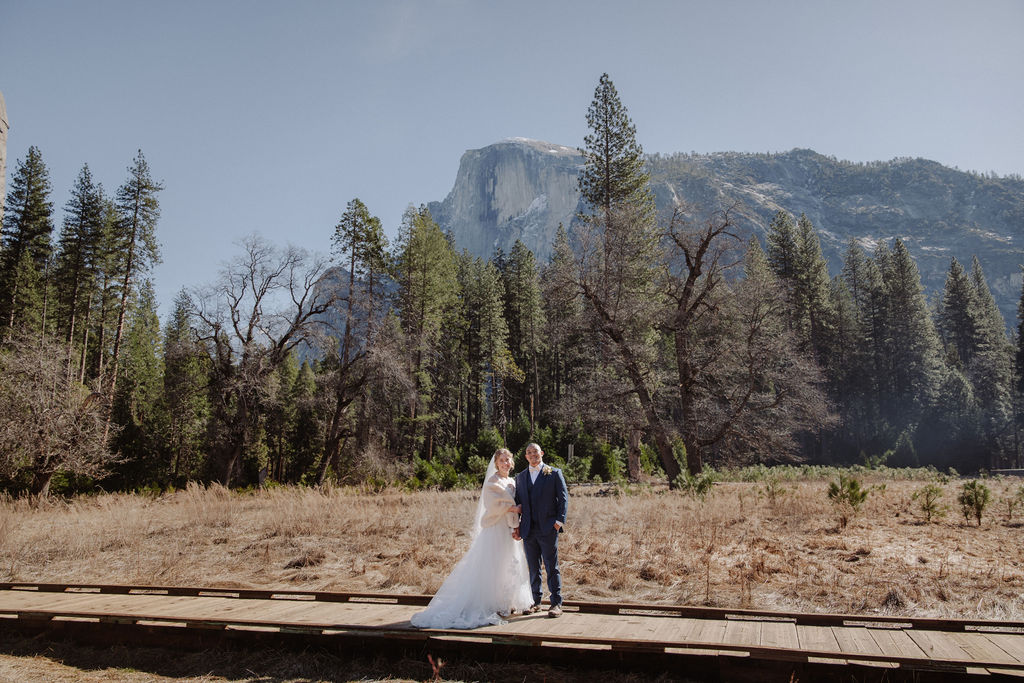 A bride and groom walk hand in hand on a paved path with tall mountains and trees in the background under a clear sky.