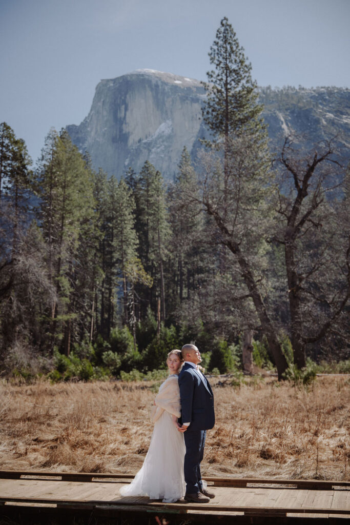 A couple in wedding attire stands outdoors on a wooden platform, with tall pine trees and a large mountain in the background.