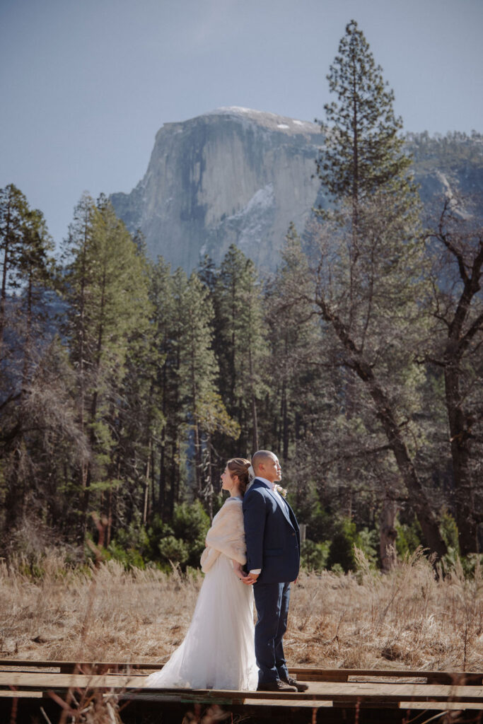 A couple in wedding attire stands outdoors on a wooden platform, with tall pine trees and a large mountain in the background.