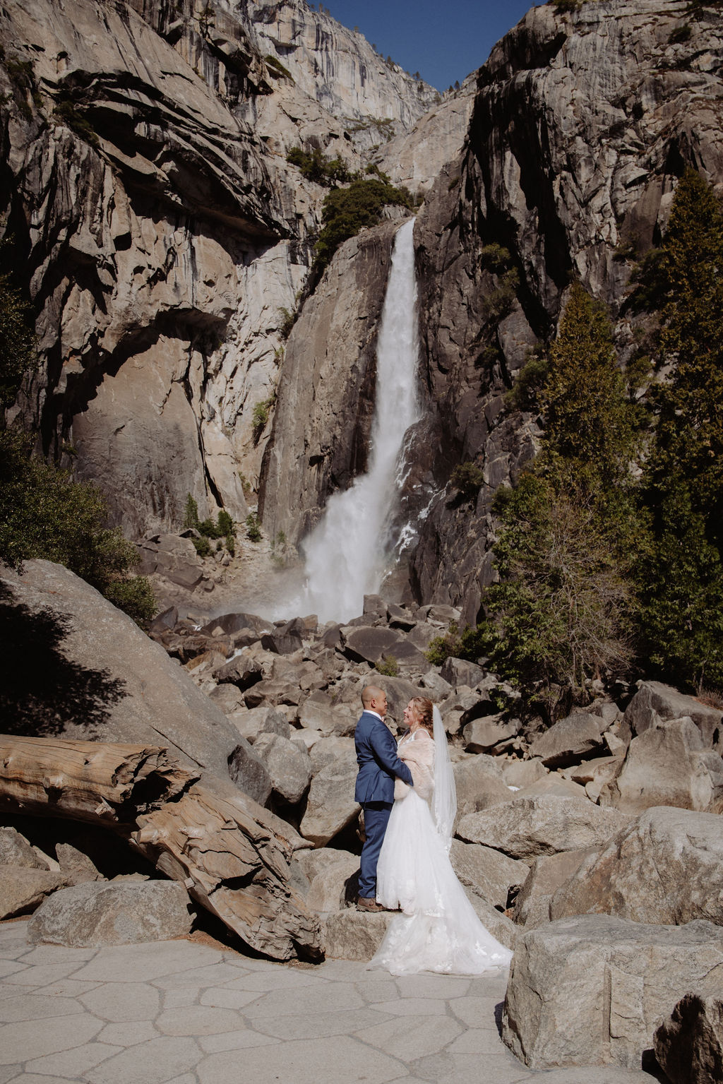 A bride and groom stand together on rocky ground in front of a tall waterfall surrounded by cliffs and trees for sunrise yosemite elopement