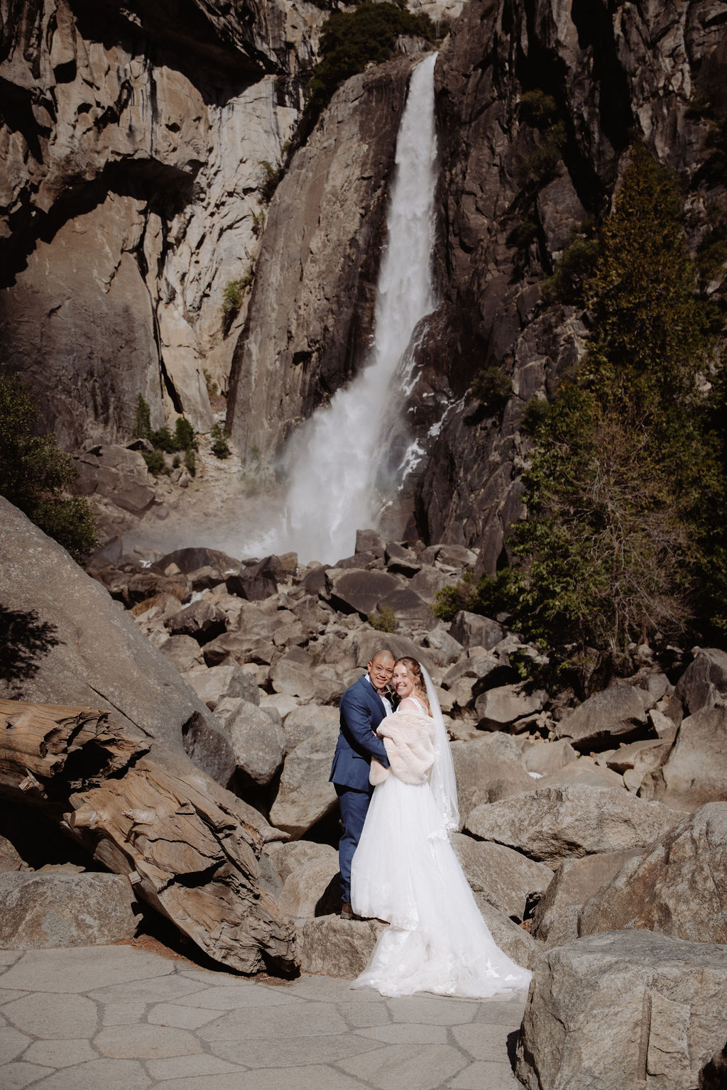A bride and groom stand together on rocky ground in front of a tall waterfall surrounded by cliffs and trees for sunrise yosemite elopement