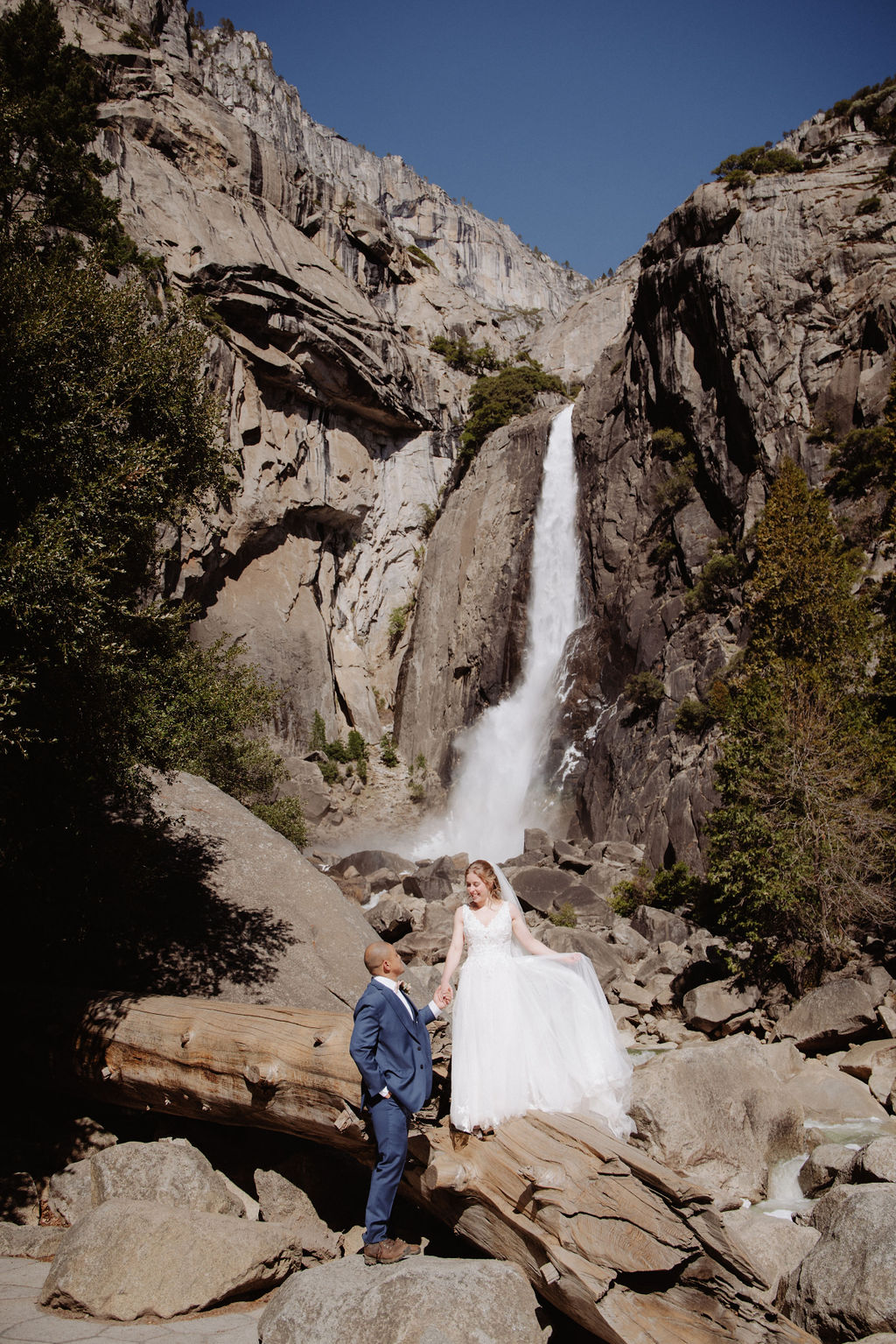 A bride and groom stand together on rocky ground in front of a tall waterfall surrounded by cliffs and trees for sunrise yosemite elopement
