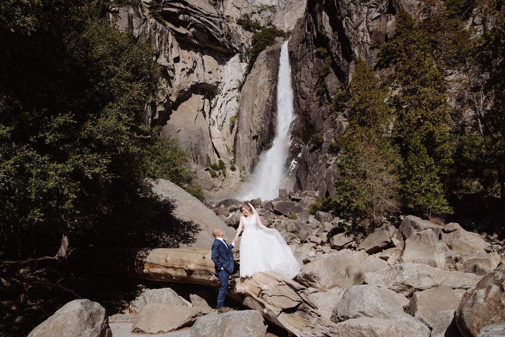 A bride and groom stand together on rocky ground in front of a tall waterfall surrounded by cliffs and trees for sunrise yosemite elopement