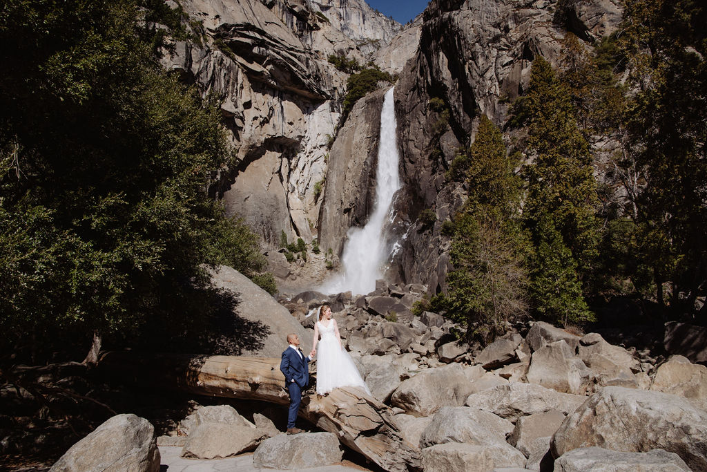 A bride and groom stand together on rocky ground in front of a tall waterfall surrounded by cliffs and trees for sunrise yosemite elopement