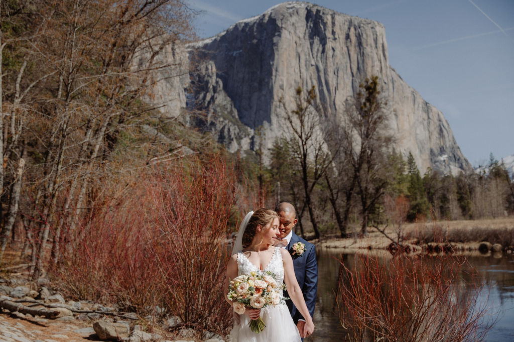 A bride and groom stand by a calm river with a rocky mountain and trees in the background on a sunny day.