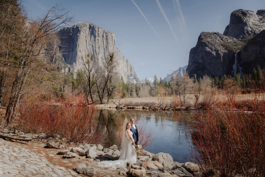 A bride and groom stand by a calm river with a rocky mountain and trees in the background on a sunny day.