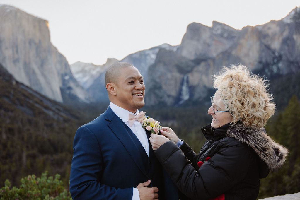 A smiling man in a navy suit gets a boutonniere pinned to his lapel by a woman, with mountains and trees in the background.