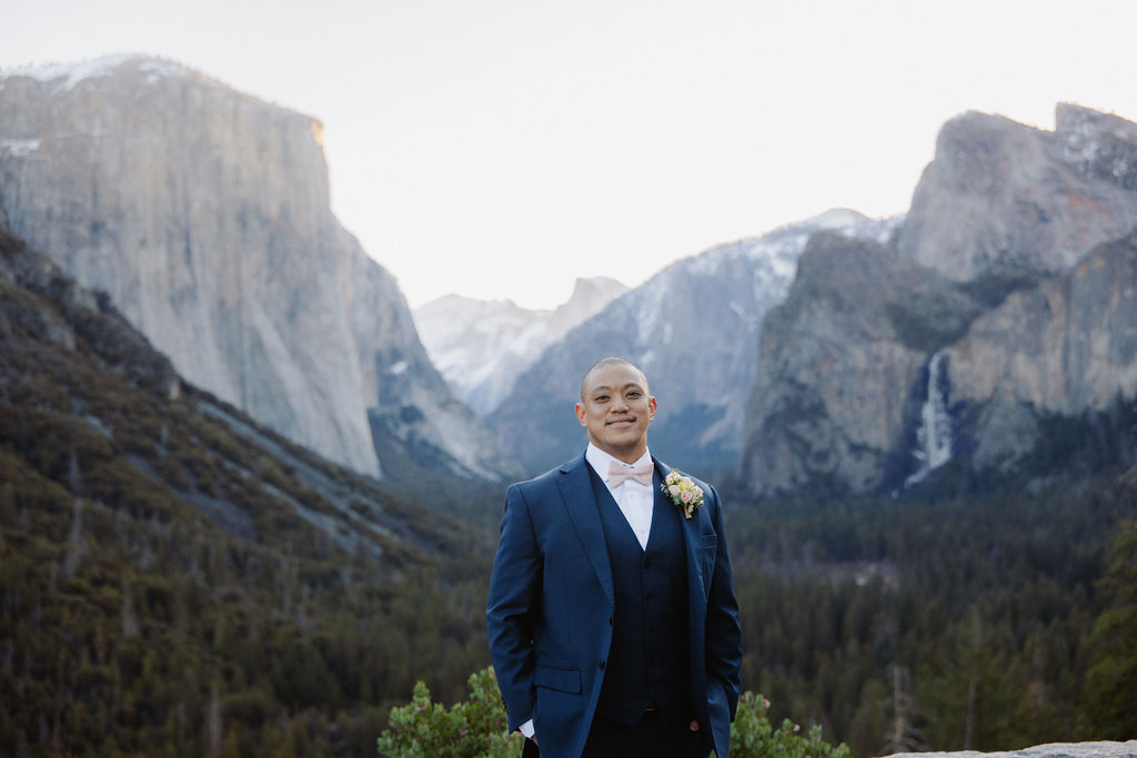 A smiling man in a navy suit gets a boutonniere pinned to his lapel by a woman, with mountains and trees in the background.