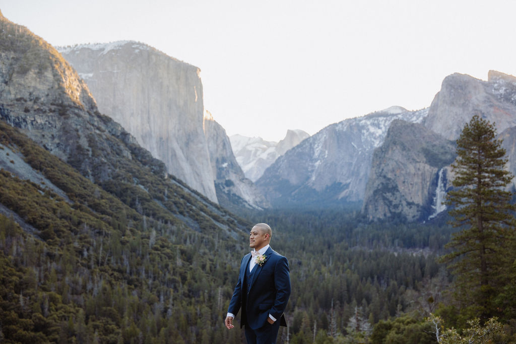 A bride and groom on a rocky ledge with mountains and forest in the background, under daylight for their sunrise yosemite elopement