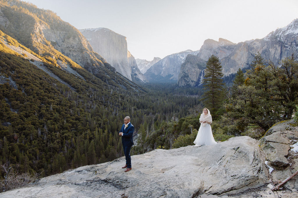A bride and groom on a rocky ledge with mountains and forest in the background, under daylight for their sunrise yosemite elopement