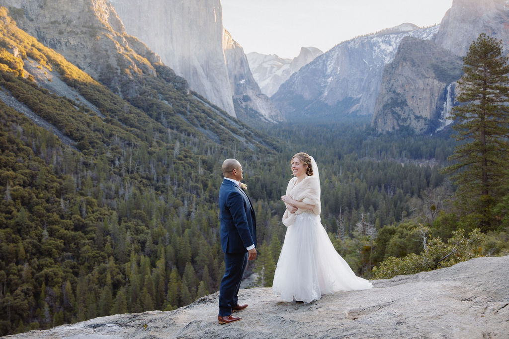 A bride and groom on a rocky ledge with mountains and forest in the background, under daylight for their sunrise yosemite elopement