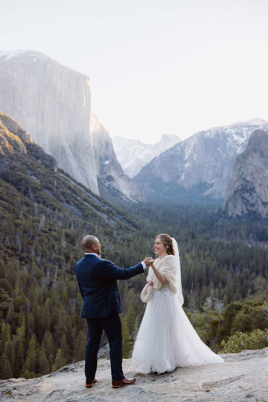 A bride and groom on a rocky ledge with mountains and forest in the background, under daylight for their sunrise yosemite elopement