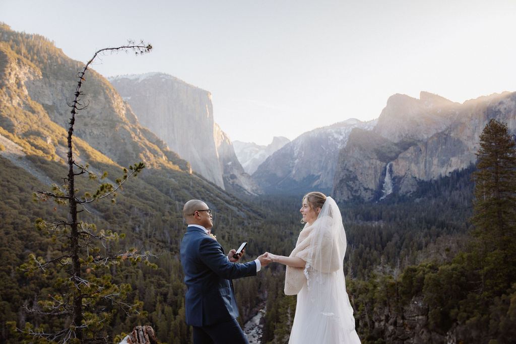 A bride and groom on a rocky ledge with mountains and forest in the background, under daylight for their sunrise yosemite elopement